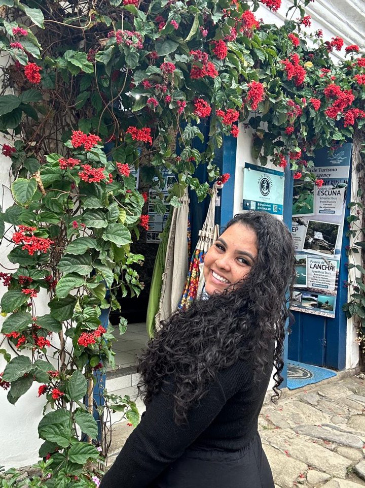 Thaiza Rangel sorrindo em frente a uma fachada com flores vermelhas